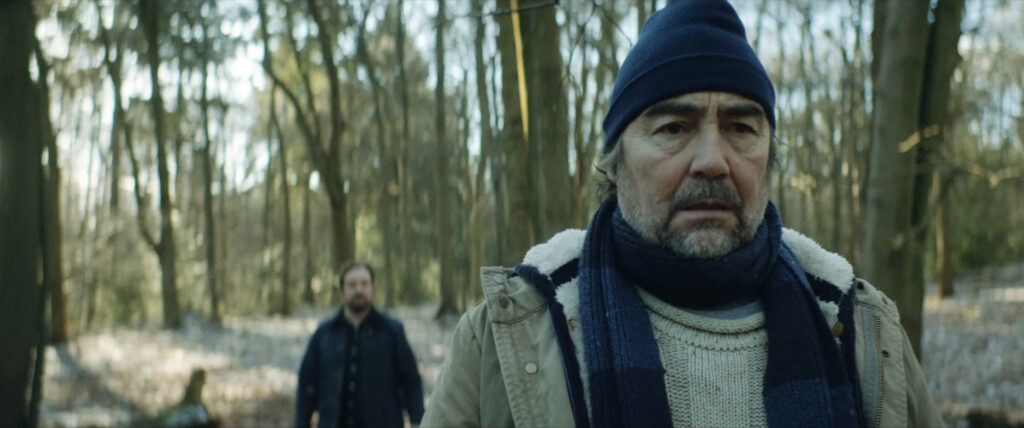 Kenny (Nathaniel Parker) in the foreground and Andy (Joel Beckett) in the background., both in the woods.