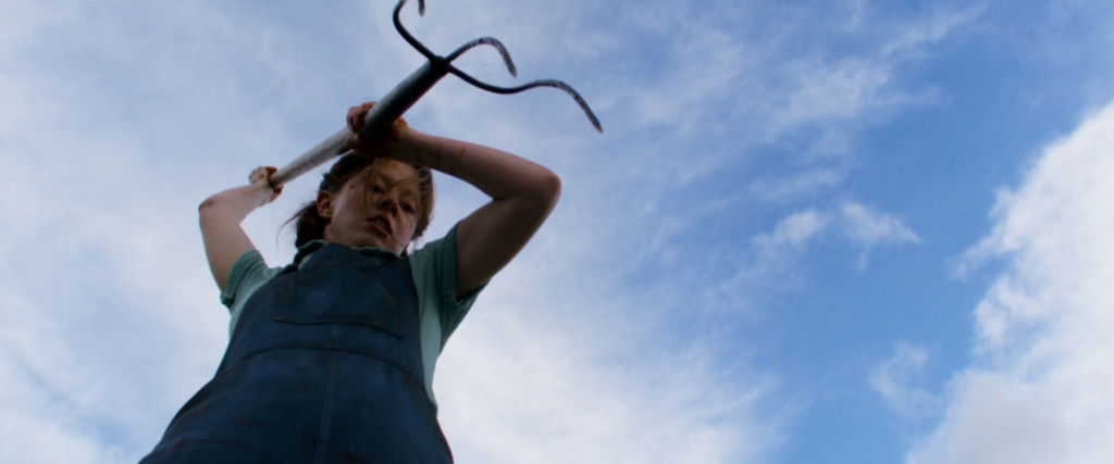 Mia Goth as Pearl, wearing blue dungarees and leveraging a pitchfork.