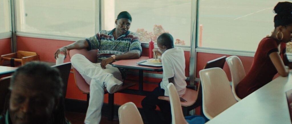 Moonlight Still; Mahershala Ali and Alex Hibbert sat in a restaurant. Ali wears white pants, floral striped shirt and a black  durag. Hibbert eats wearing a white tshirt and black trousers.