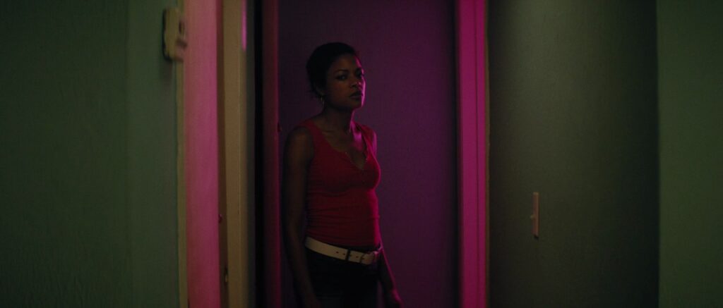 Naomie Harris in Moonlight stood in  a purple-tinted doorway, wearing a sleeveless pink top, white belt and jeans