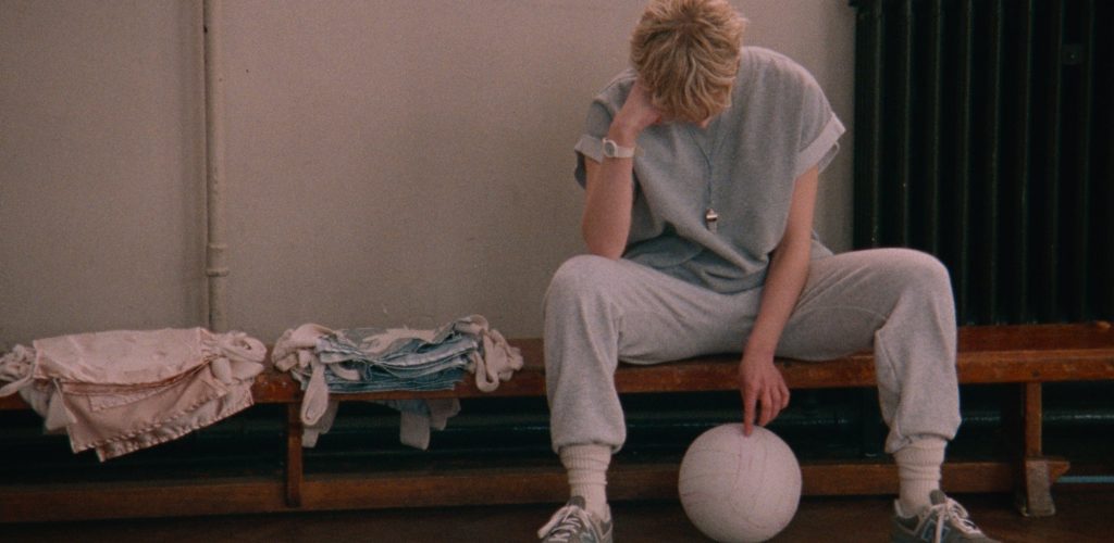 Rosy McEwen (blonde white woman wearing grey sweatpants and tshirt) sits on a gym bench next to bibs and poking at a netball.